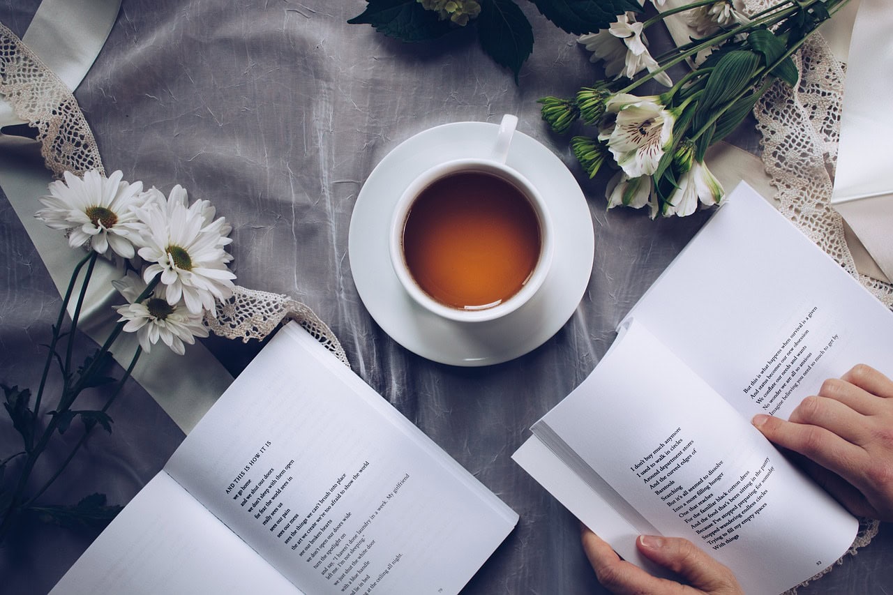 A cup of tea in a white tea cup on a grey cloth surrounded by white flowers and two open books